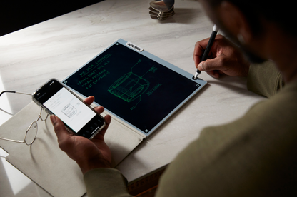 Person using a stylus on a dark-mode smart notebook with a smartphone nearby on a light-colored surface.